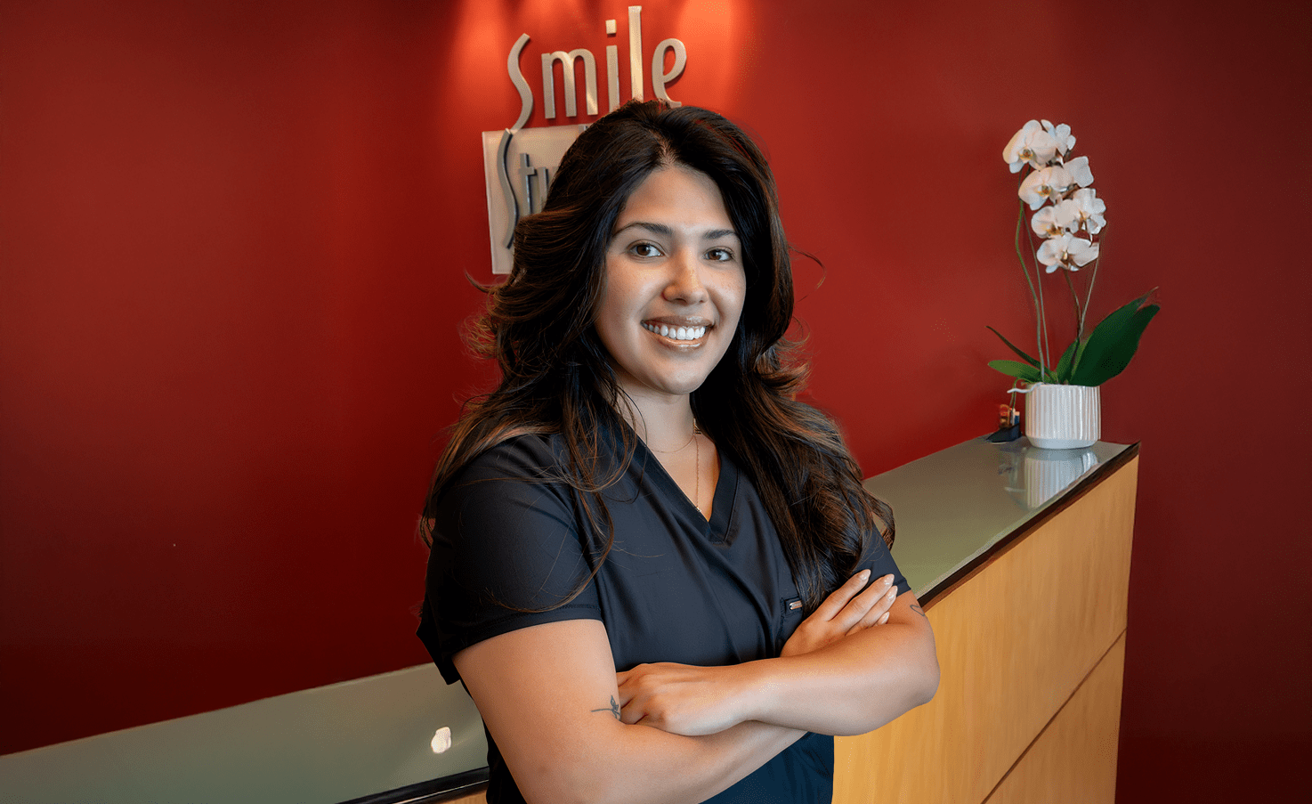 A woman in a black uniform smiles confidently at the camera, standing in front of a red wall with "Smile" visible. A potted orchid adds elegance.