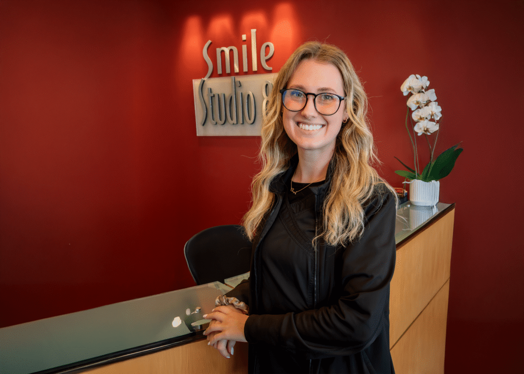 A smiling person with glasses stands at a dental office reception desk, under a sign reading "Smile Studio." The background is red with a potted white orchid, conveying a welcoming atmosphere.