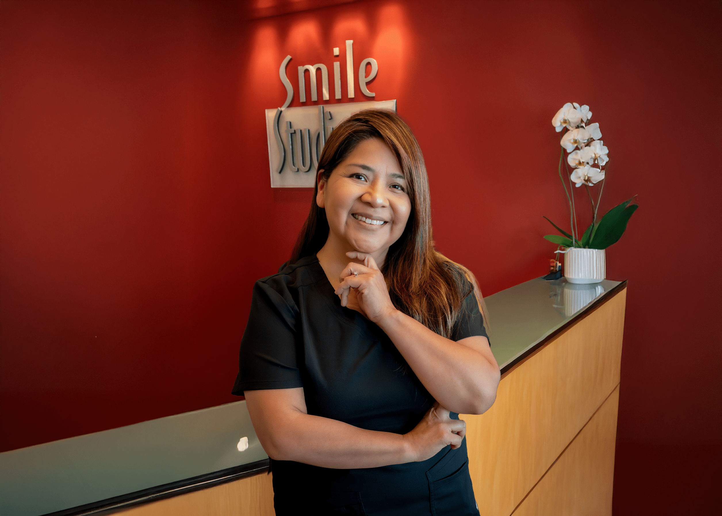 A woman in a black outfit smiles warmly at a reception desk against a red wall with "Smile Studio" signage. An orchid adds a touch of elegance.
