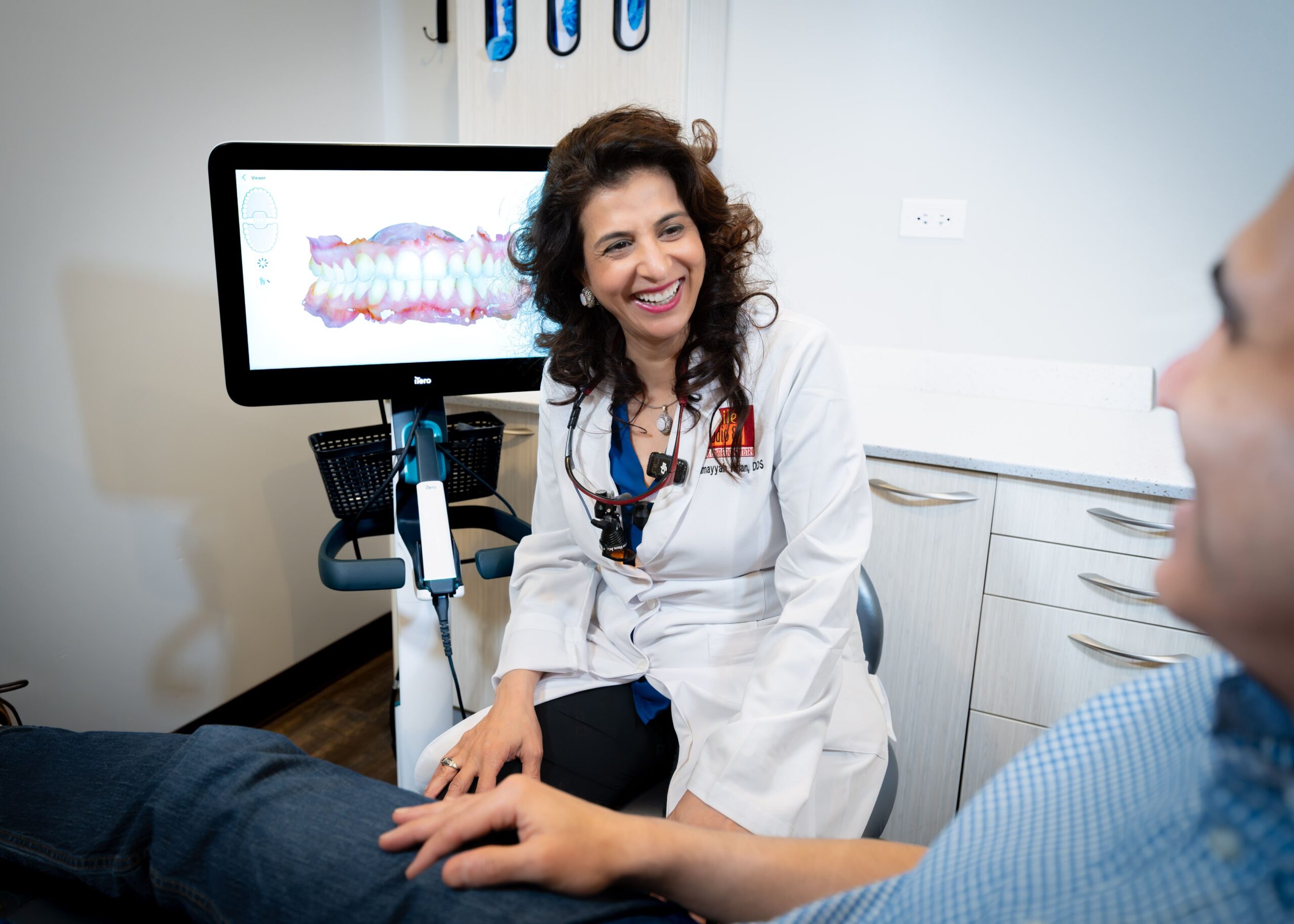 Dentist in a white coat smiles warmly at a seated patient. A screen showing a 3D dental scan is visible. The room is modern and bright.
