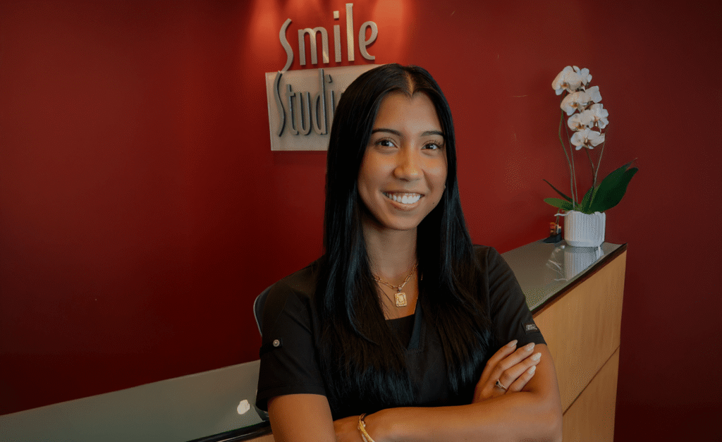 A woman with long dark hair smiles in a dental office lobby, standing with arms crossed. Behind her, a red wall displays "Smile Studio" and a white orchid.
