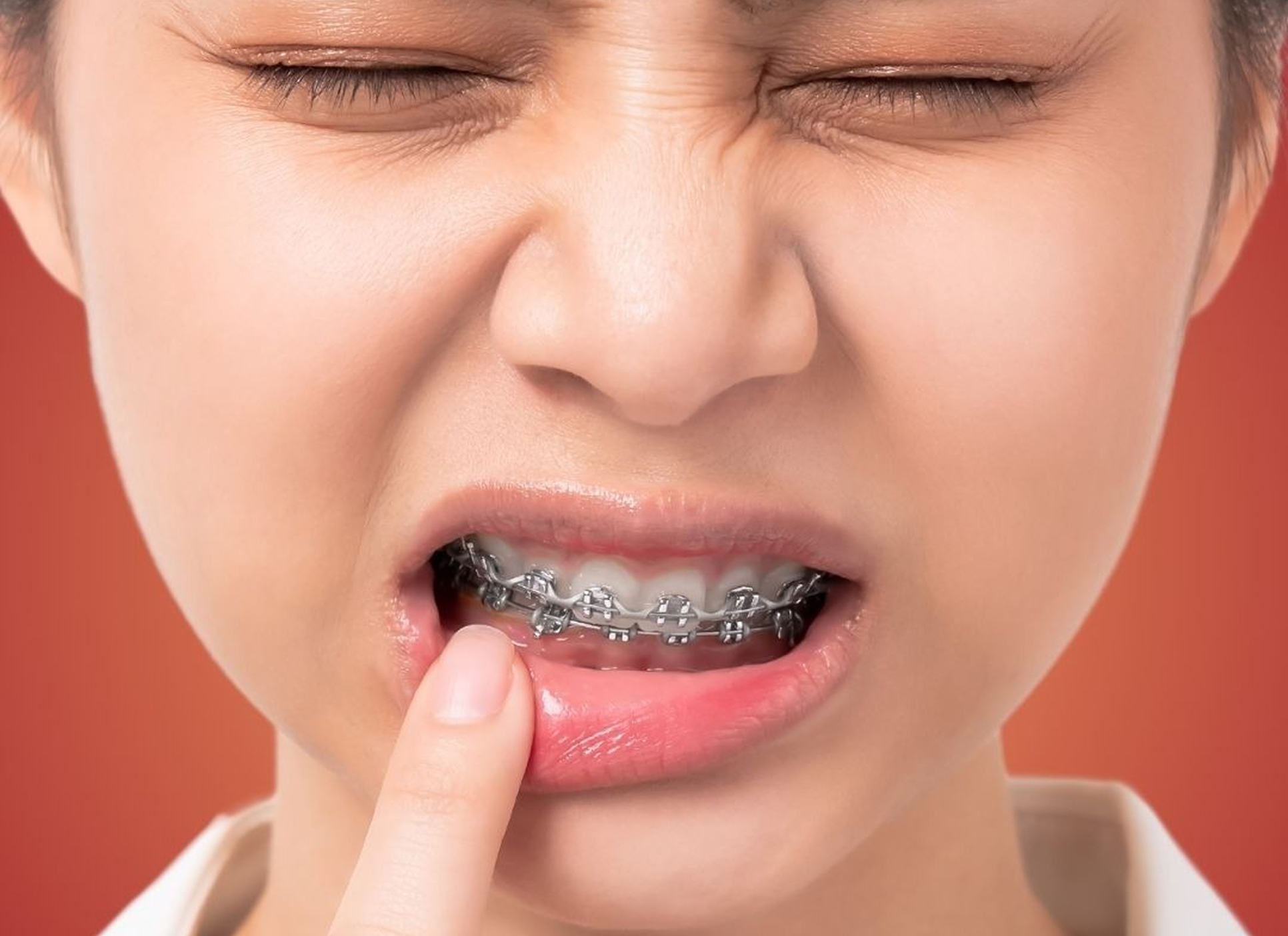 A person with braces winces in discomfort, eyes closed, and touches their lower lip with a finger against an orange background, conveying dental pain.