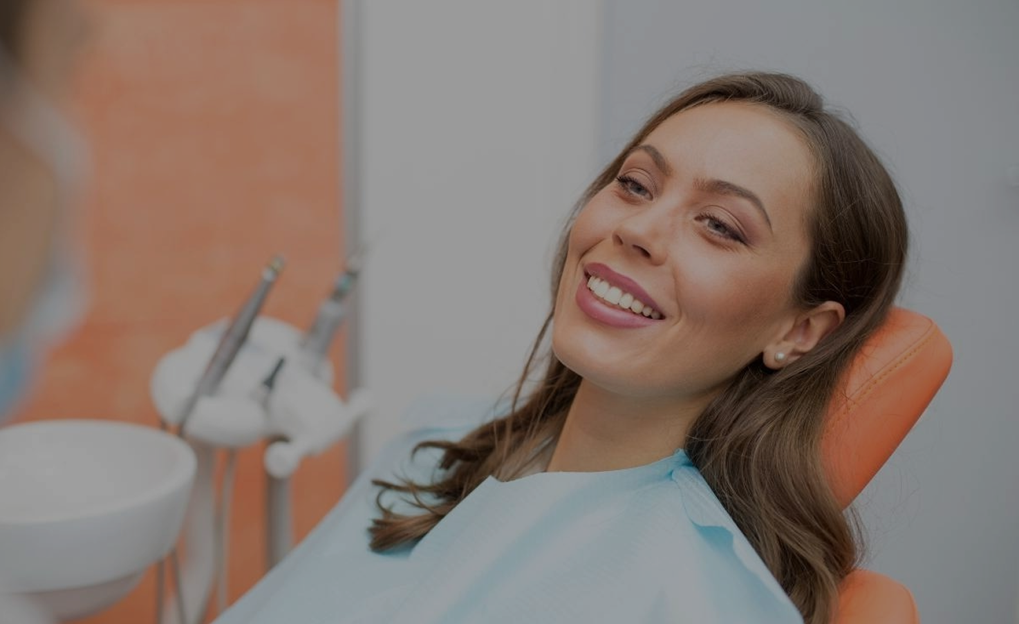 Smiling woman in a dental chair, wearing a blue bib. Dental tools and orange wall in the background, conveying a calm, positive atmosphere.