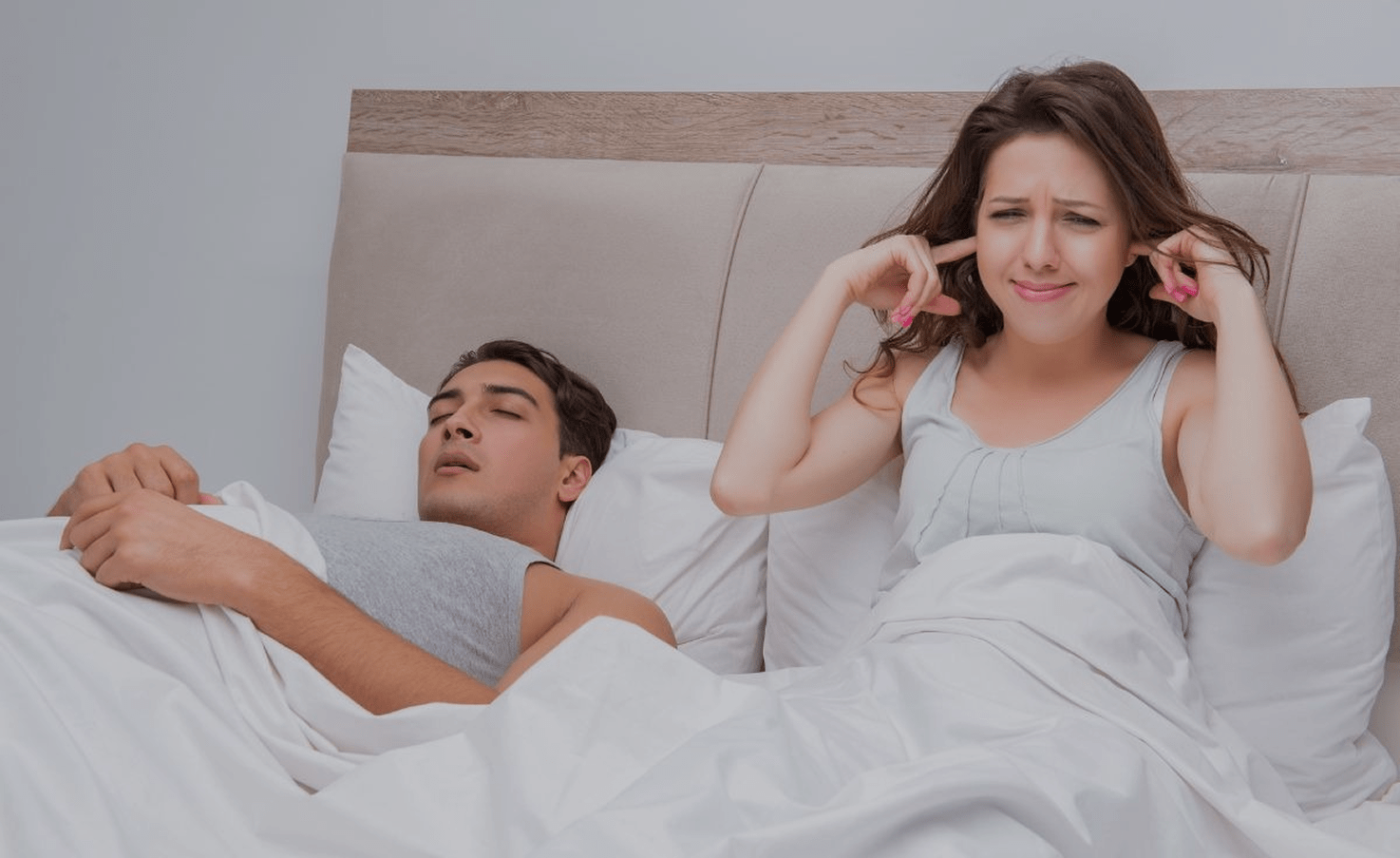 A woman in bed covers her ears in frustration while a man sleeps peacefully beside her, suggesting he is snoring. The mood is humorous and relatable.