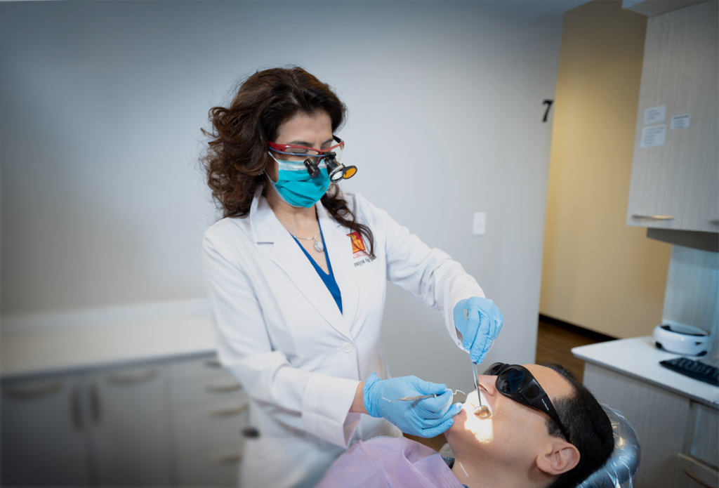 A dentist in a white coat and magnifying glasses examines a patient's teeth, who is wearing protective eyewear. The setting is a modern dental office.