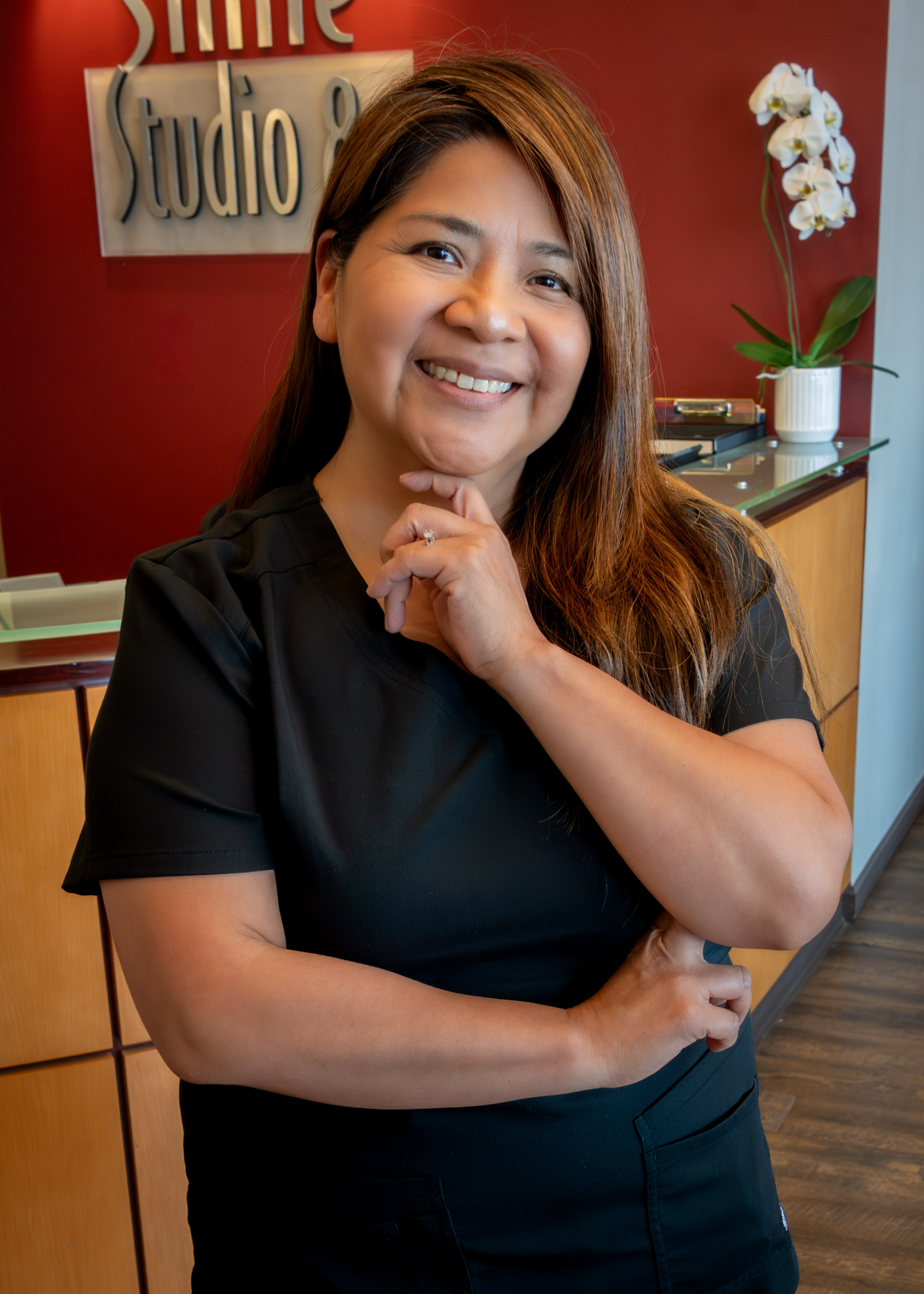A woman in a black outfit smiles warmly, standing in an office with red walls. A sign behind her reads "Smile Studio 8," and there is a white orchid on the counter.