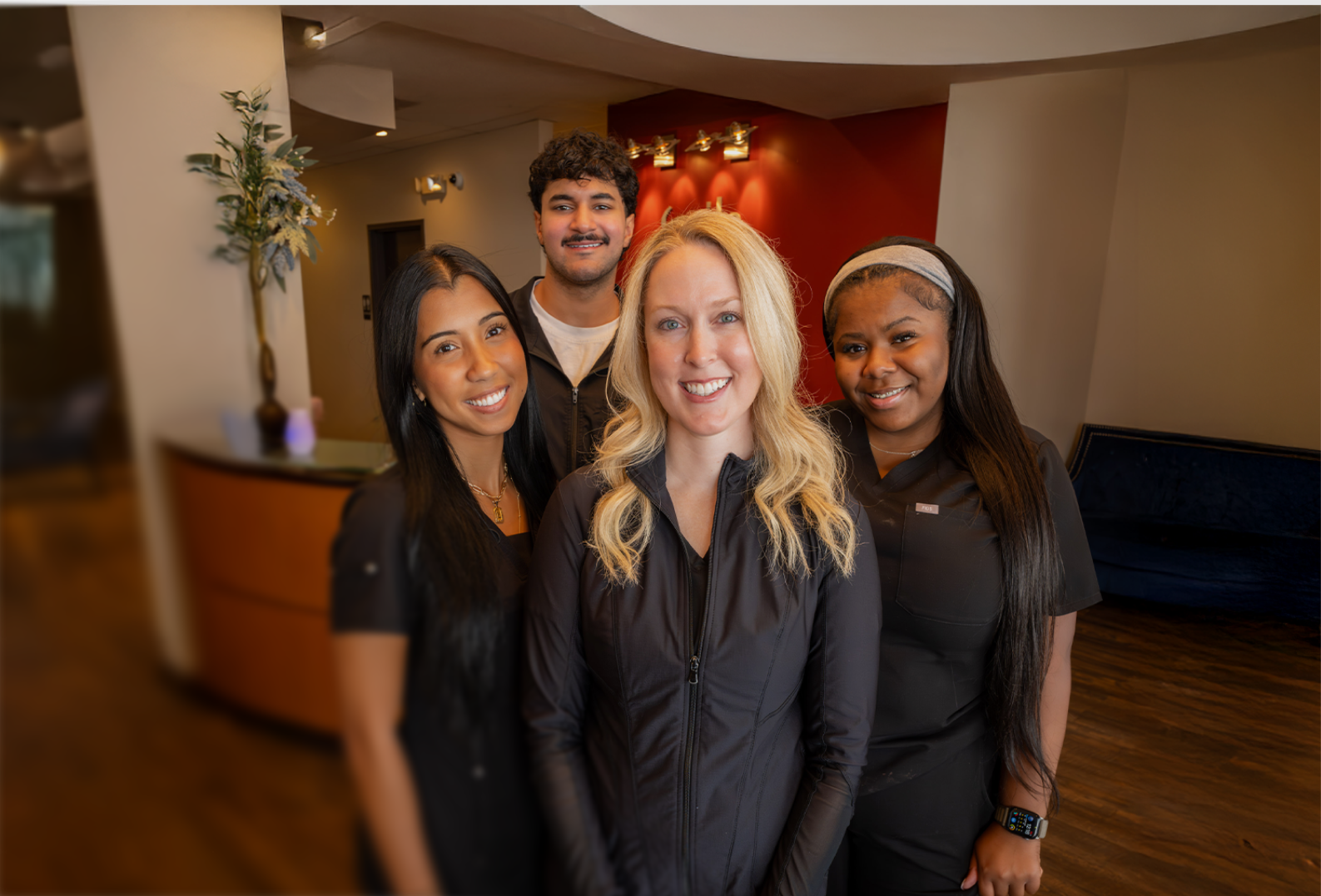 A group of four smiling individuals stands in a warmly lit office lobby with a red accent wall, wooden floor, and a potted plant, conveying a welcoming atmosphere.