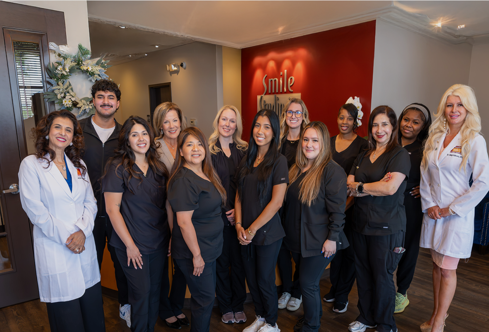 A group of eleven dental professionals, mainly in black scrubs, pose smiling in an office with "Smile" on the red wall, conveying a warm atmosphere.