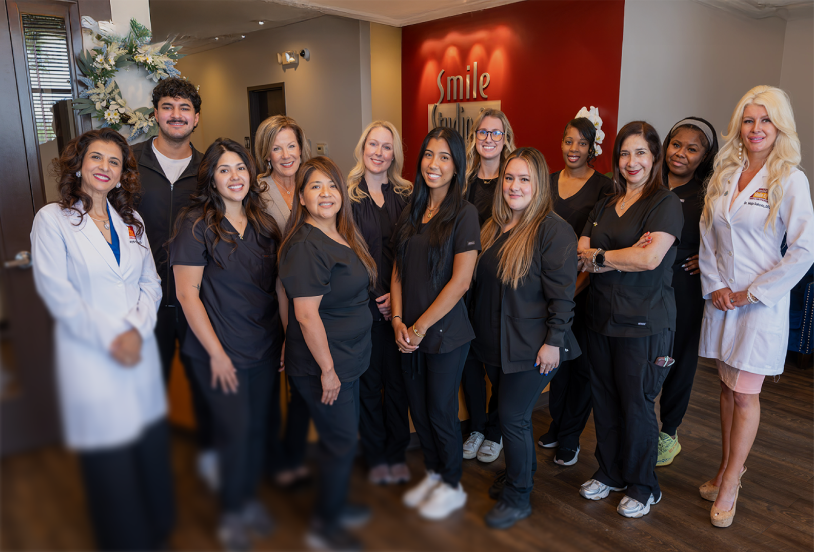 A group of eleven dental professionals, mainly in black scrubs, pose smiling in an office with "Smile" on the red wall, conveying a warm atmosphere.